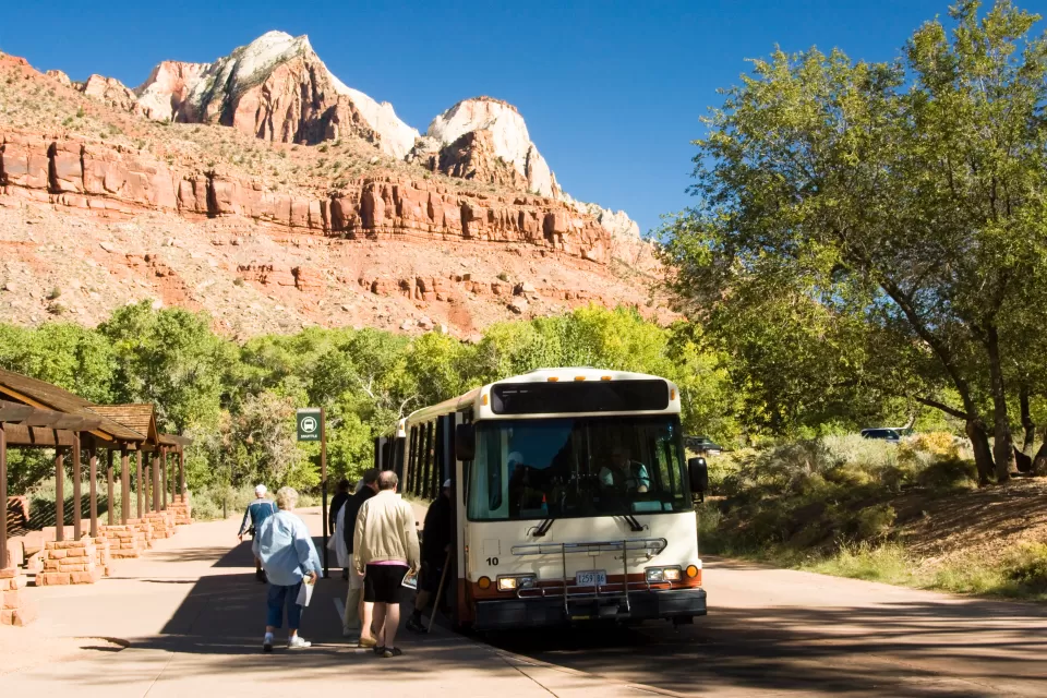 White shuttle bus picks up passengers in Zion National Park, red cliffs and mountains in background.